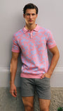 Man wearing a pink patterned polo shirt and gray shorts standing against a plain wall.