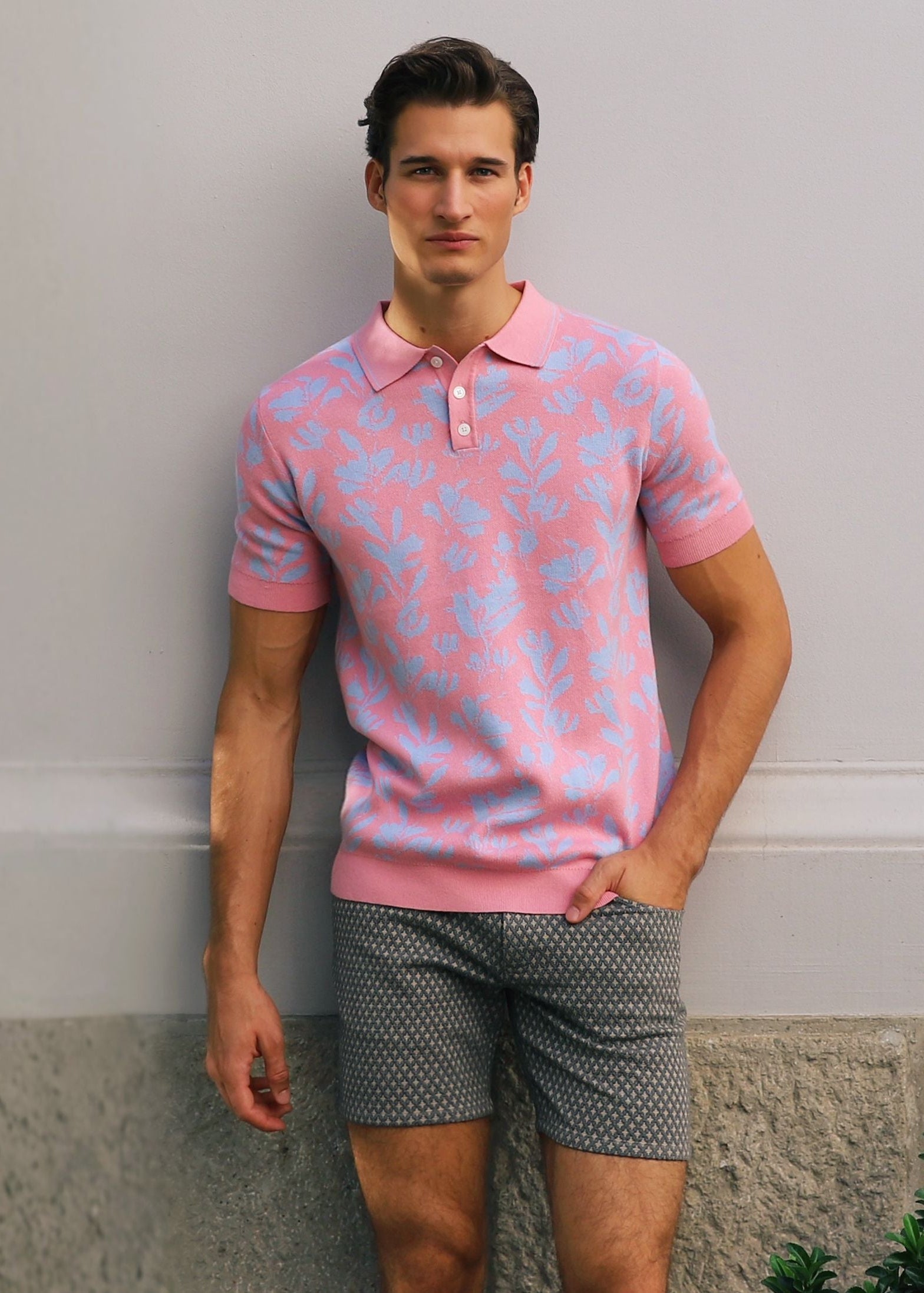 Man wearing a pink patterned polo shirt and gray shorts standing against a plain wall.