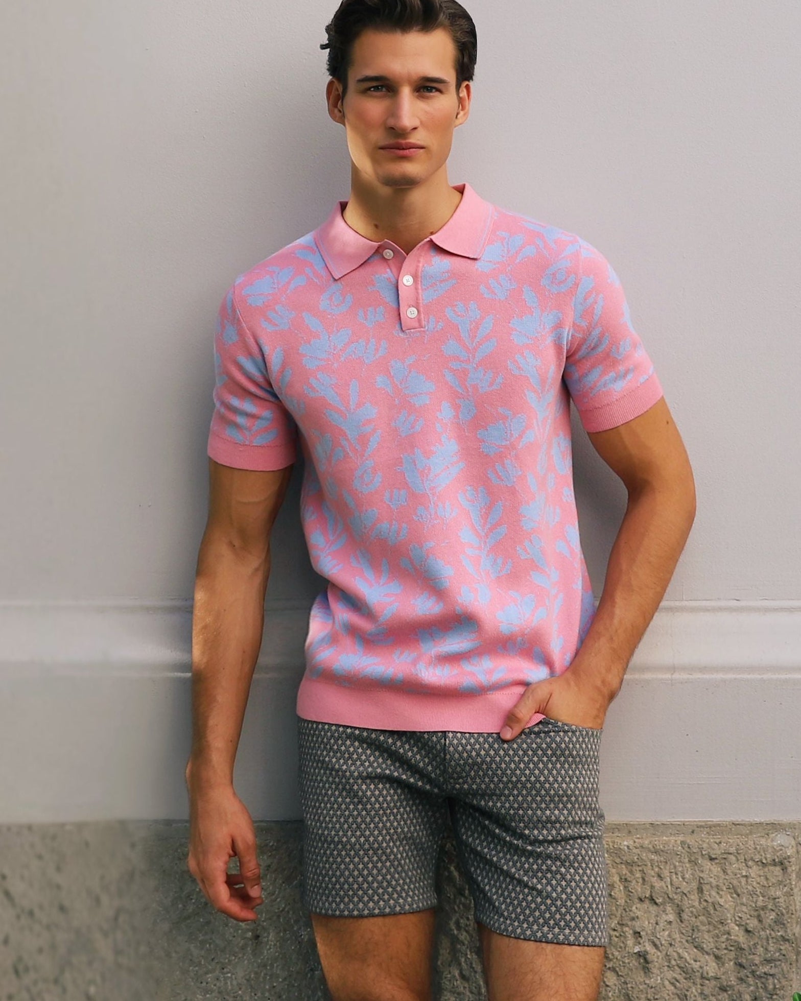 Man wearing a pink patterned polo shirt and gray shorts standing against a plain wall.