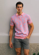 Man wearing a pink patterned polo shirt and gray shorts standing against a plain wall.