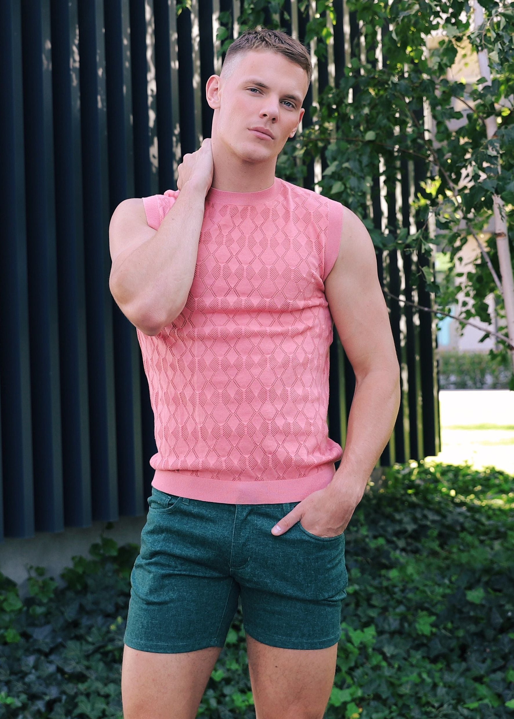 Man wearing a pink sleeveless shirt and green shorts standing outdoors.