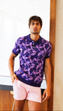 Man wearing a purple patterned shirt and pink shorts standing in a room with wooden floor and white walls.