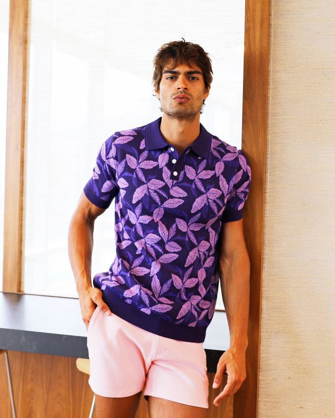 Man wearing a purple patterned shirt and pink shorts standing in a room with wooden floor and white walls.