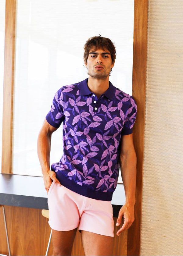 Man wearing a purple patterned shirt and pink shorts standing in a room with wooden floor and white walls.