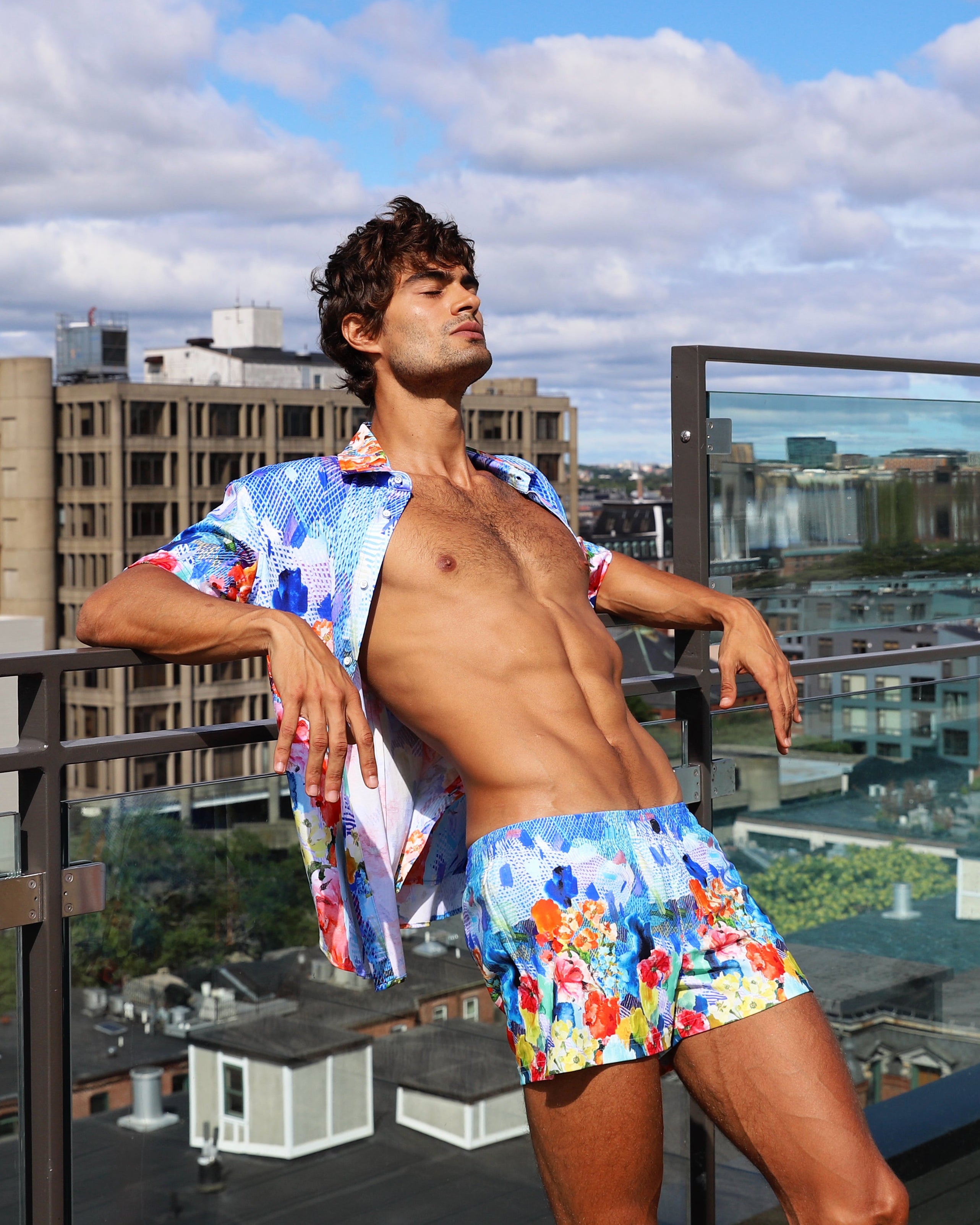Man in colorful swim trunks standing on a rooftop with cityscape in the background