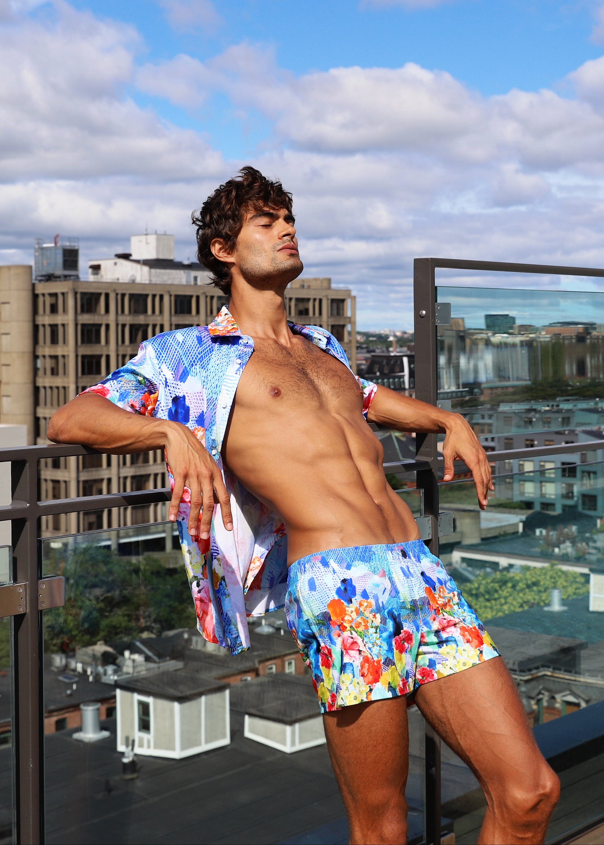 Man in colorful swim trunks standing on a rooftop with cityscape in the background