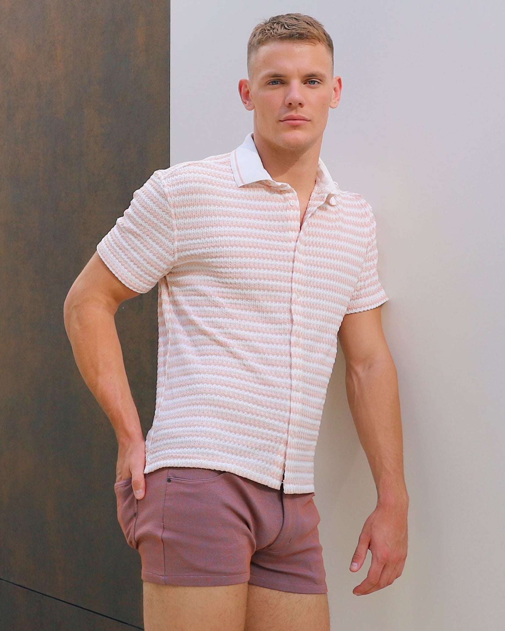 Man wearing a striped shirt and shorts standing against a textured wall.