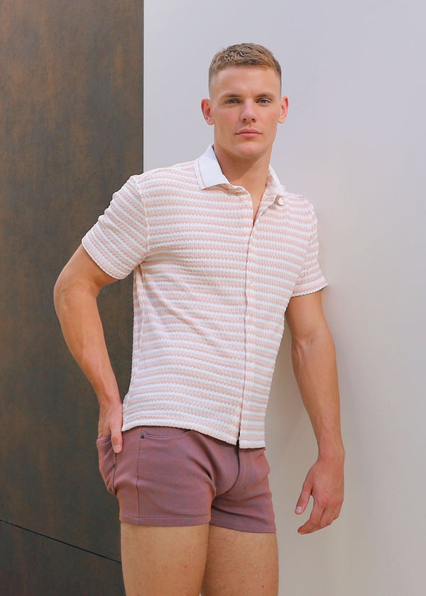 Man wearing a striped shirt and shorts standing against a textured wall.