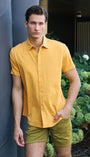 Man in a yellow shirt and green shorts standing outdoors near a building with greenery.