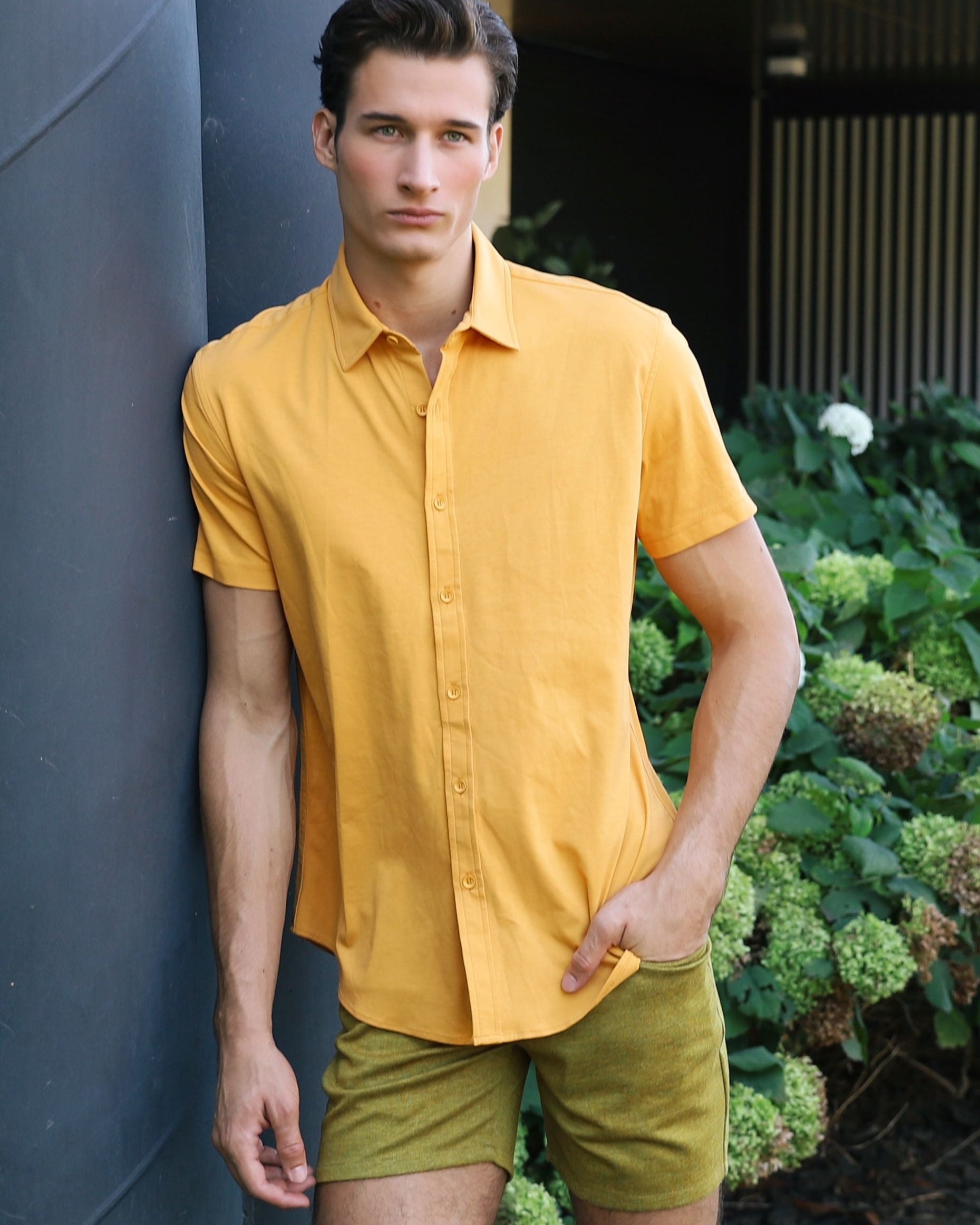Man in a yellow shirt and green shorts standing outdoors near a building with greenery.