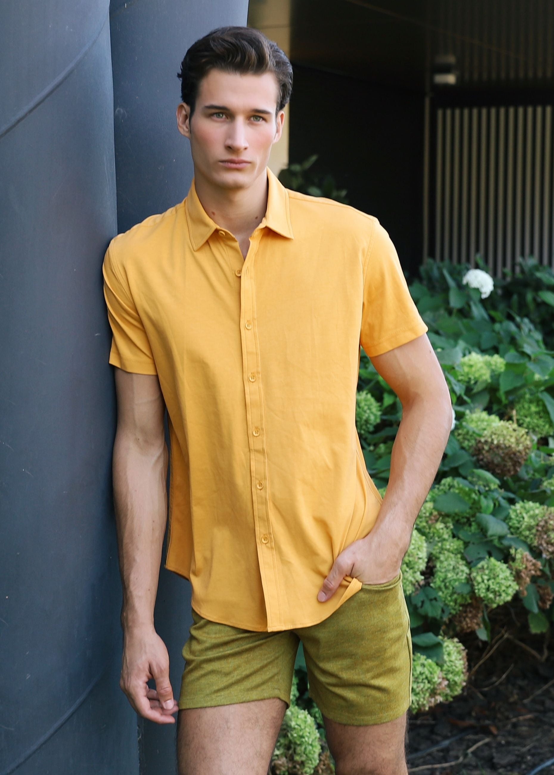 Man in a yellow shirt and green shorts standing outdoors near a building with greenery.