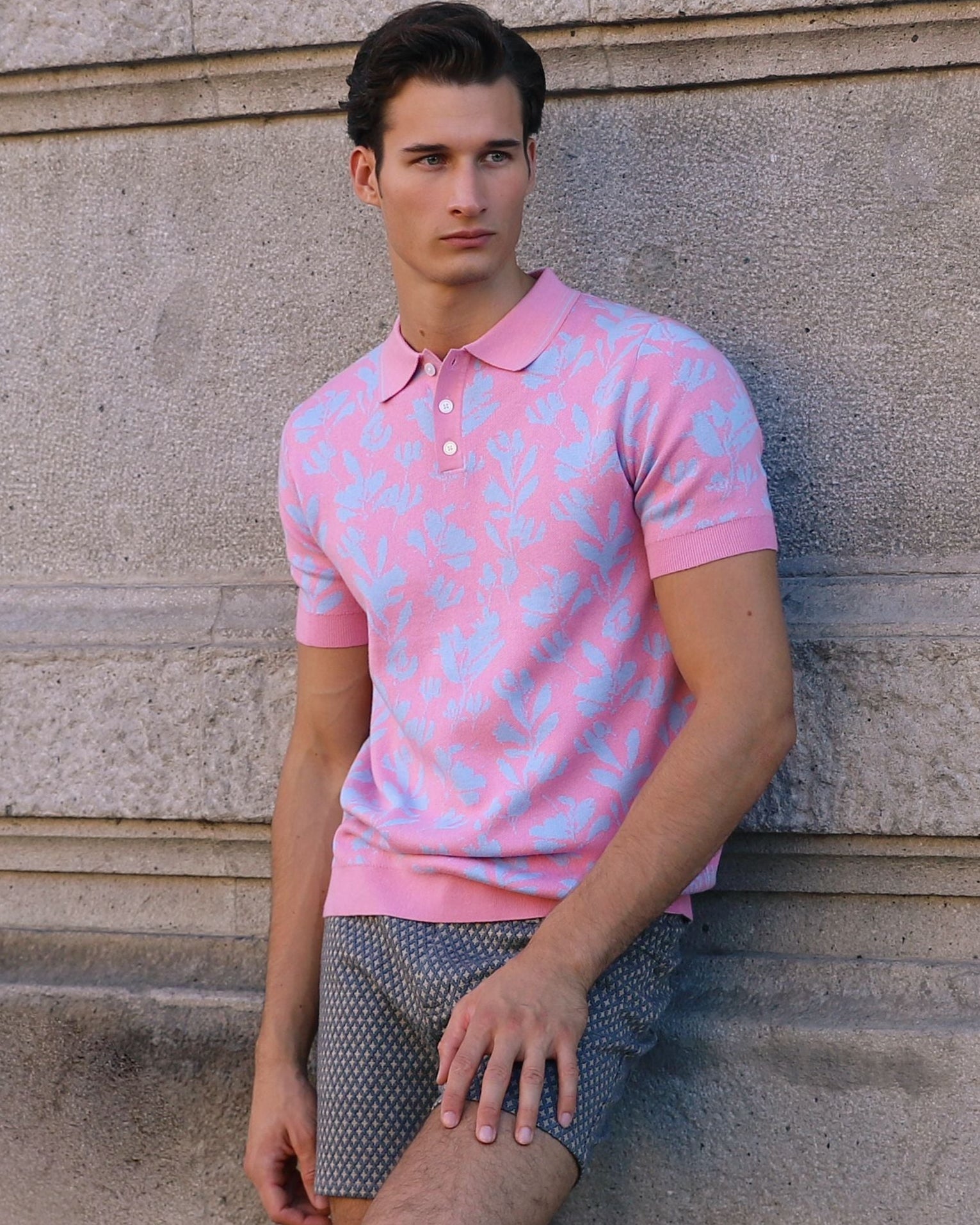 Man wearing a pink polo shirt leaning against a stone wall.