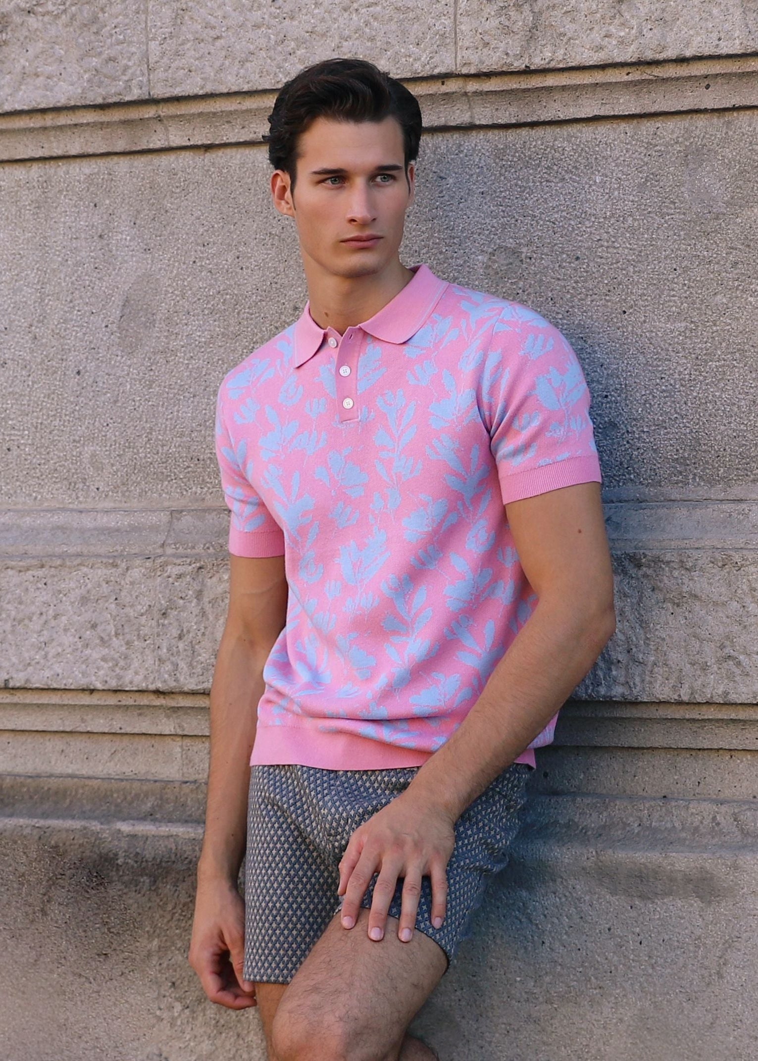 Man wearing a pink polo shirt leaning against a stone wall.
